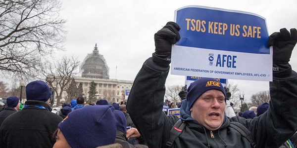 Image of man holding a rally sign that says TSOS Keep Us Safe at a rally outside the US Capitol building.