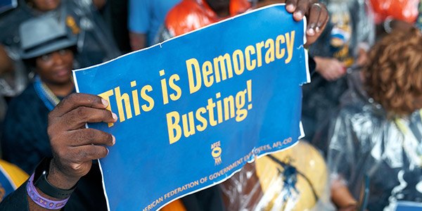 Close up of rally sign with the text This is Democracy Busting! Rally crowd is in the background. 