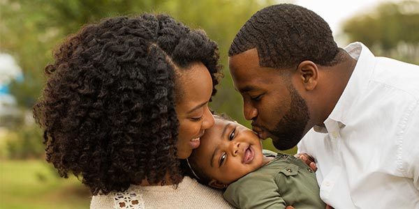 A Black woman and man hold a child together while kissing the child's face. The child is smiling. 