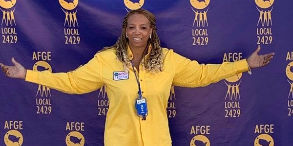 Image of Roslyn Austin-Stewart. She is wearing a yellow long sleeve shirt standing in front of a blue backdrop with the all gold AFGE logo repeating.