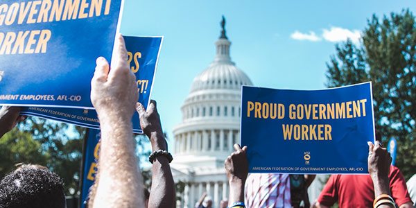 Rally goer holds above their head with the Capitol building in the background. The sign reads Proud Government Worker