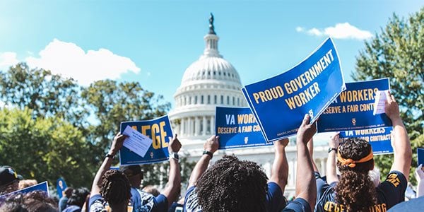 Rally-goer in the foreground holding sign that says Proud Government Worker. US Capitol Building can be seen in the background.