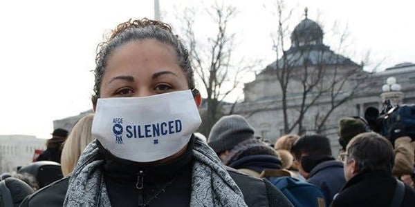 Woman standing outside at a rally with a mask covering the lower part of her face that says Silenced. 