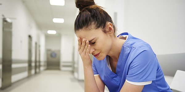 Woman nurse in blue scrubs sits in hospital hallway holding a hand to her forehead while leaning forward. 