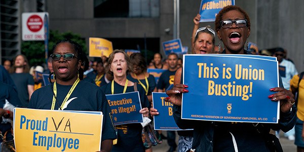 Two women in the foreground at a rally. One is holding a sign that says "This is Union Busting! The other says "Proud VA Employee"