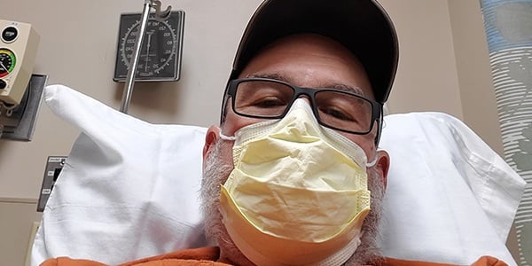 Close up of man wearing glasses and a cap with a mask over his nose and mouth, laying in a hospital bed. 