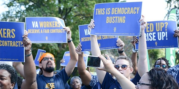 Image of rally goers holding signs outside of the U.S. Capitol. some of the signs say This is Democracy Busting and VA Workers Deserve a Fair Contract