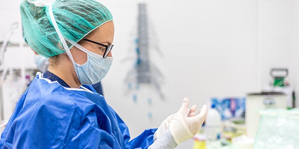 Healthcare worker wearing medical gown, glasses, and hair protector is putting on gloves