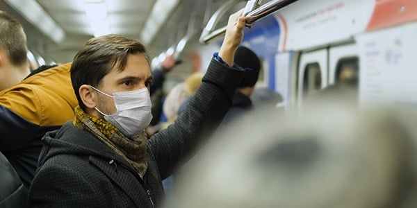 Image of man holding overhead train rail on crowded train. They are wearing a mask.