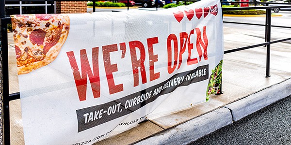 Image of a We're Open banner hanging from a black railing on a side walk. The banner has pizza images in the corners. 