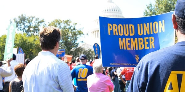 Image of backs of individuals attending a rally in front of the US Capitol. In the foreground is a person holding a blue and gold sign that says Proud Union Member