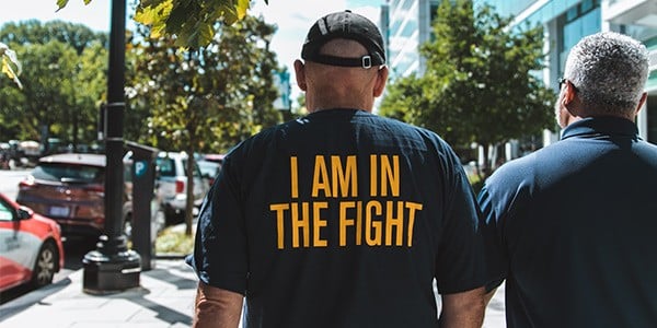 Image of the back of two people walking down a sidewalk. The back of one person's shirt says I Am In The Fight