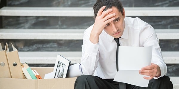 White man sitting on steps with his hand on his forehand and holding a sheet of paper. A box of office items is beside him.