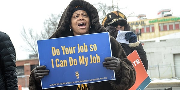 Black woman wearing a coat holding a sign at a rally that reads Do Your Job So I Can Do My Job