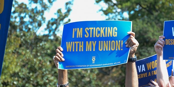 Two arms holding up a blue and gold rally sign that says I'm Sticking With My Union