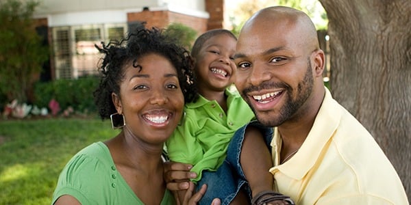 Black family with woman, man and childing standing in front of house smiling.