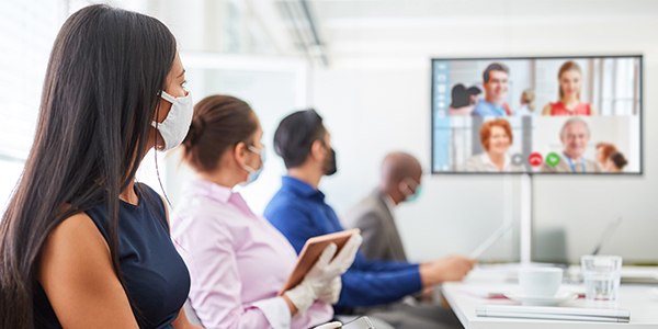 Workers wearing masks sitting in a conference room with a virtual meeting on the TV screen